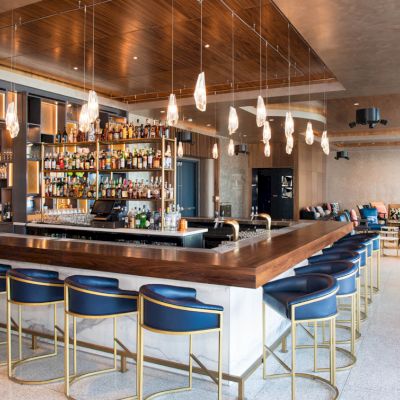A modern bar with a large selection of bottles, sleek bar stools, pendant lights, and a cozy seating area in a stylish space.