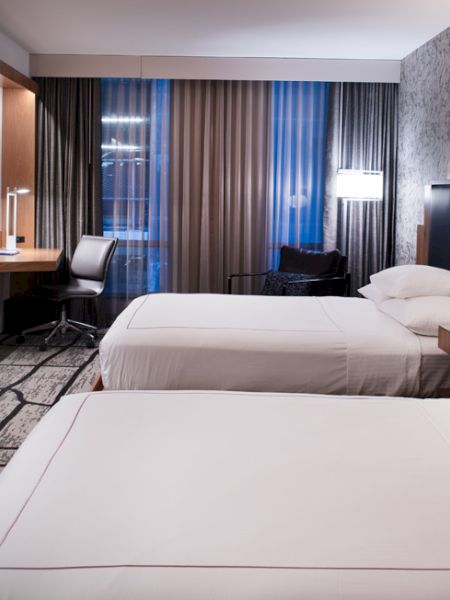 A hotel room with two beds, a TV, desk, chair, and modern decor, featuring patterned walls and curtains creating a cozy atmosphere.