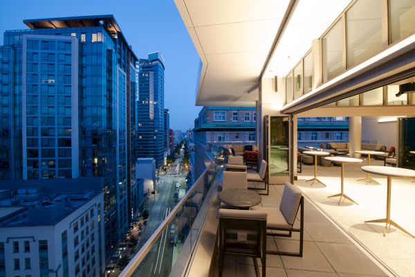 A modern cityscape at dusk from a high-rise balcony with sleek furniture, overlooking illuminated streets and tall buildings.