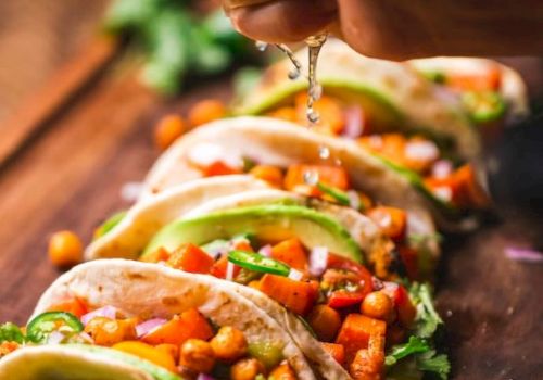 A person squeezes lime over a row of tacos filled with vegetables and chickpeas on a wooden board, garnished with cilantro and lime slices.