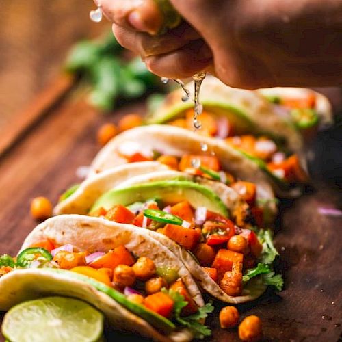 A person squeezes lime over a row of tacos filled with vegetables and chickpeas on a wooden board, garnished with cilantro and lime slices.