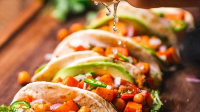 A person squeezes lime over a row of tacos filled with vegetables and chickpeas on a wooden board, garnished with cilantro and lime slices.