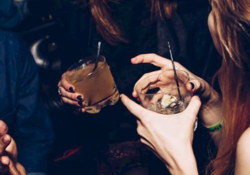 People are enjoying drinks and laughing together in a lively setting, with two women holding cocktail glasses and someone gesturing.