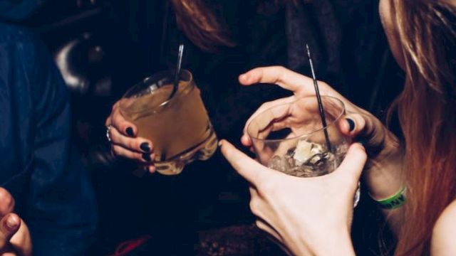 People are enjoying drinks and laughing together in a lively setting, with two women holding cocktail glasses and someone gesturing.