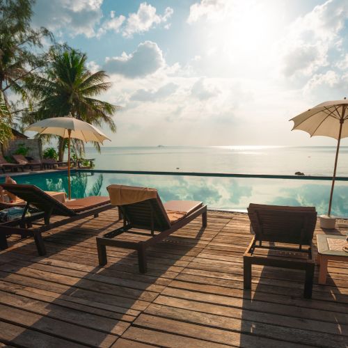 Lounge chairs and umbrellas by a poolside with a view of the ocean under a partly cloudy sky at sunset.