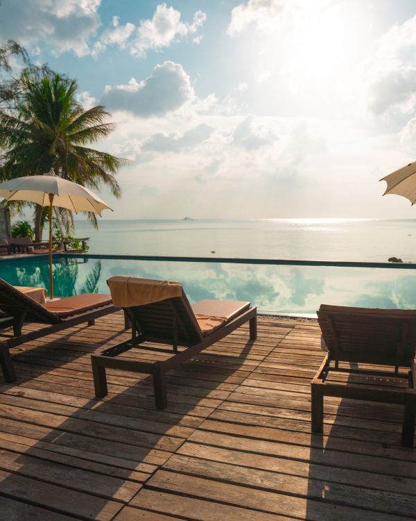 Lounge chairs and umbrellas by a poolside with a view of the ocean under a partly cloudy sky at sunset.