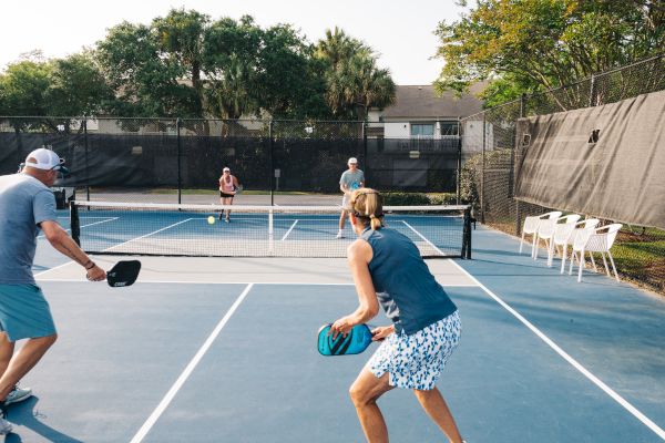 People playing pickleball on an outdoor court with trees and chairs nearby.
