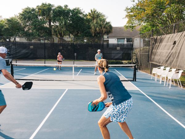 People playing pickleball on an outdoor court with trees and chairs nearby.