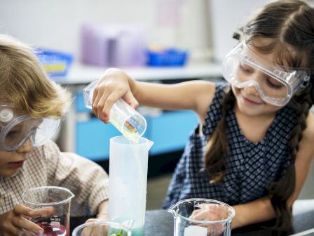 Two children wearing safety goggles engage in a science experiment, pouring liquid from a beaker into a measuring cylinder.