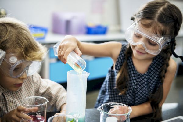 Two children wearing safety goggles engage in a science experiment, pouring liquid from a beaker into a measuring cylinder.
