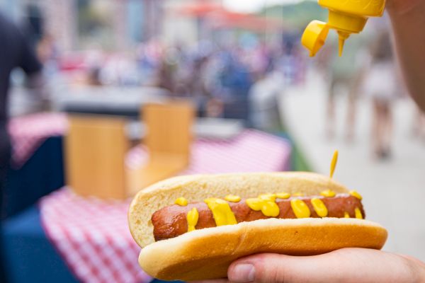 A person is squeezing mustard onto a hot dog held in one hand at an outdoor setting with tables in the background.