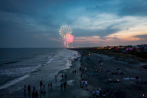 A beach at dusk with groups of people, under a sky lit by colorful fireworks.