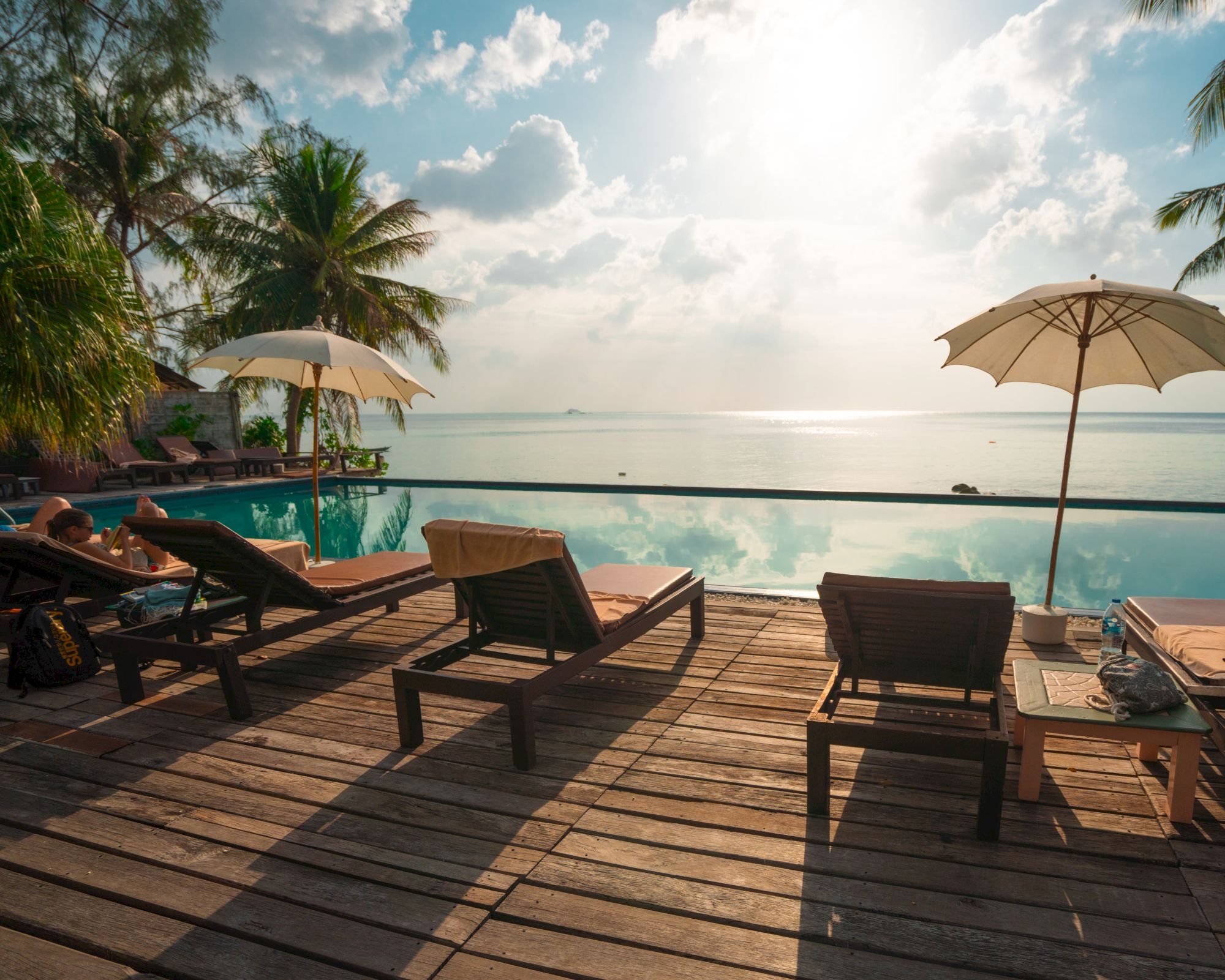 A serene poolside with lounge chairs, umbrellas, and a view of palm trees and the ocean at sunrise or sunset.