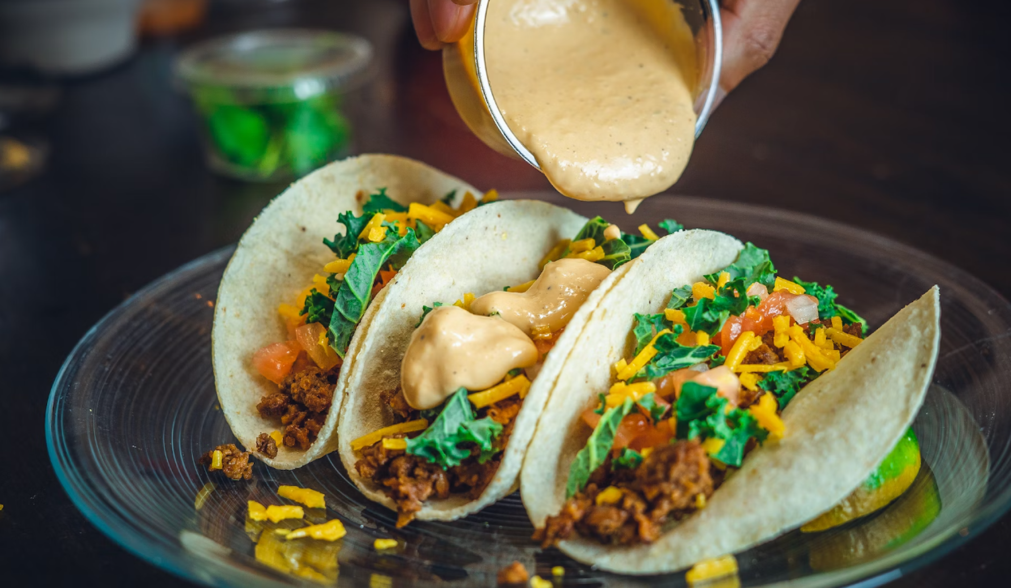 Three tacos with toppings like cheese and greens are on a plate as sauce is being poured over them.