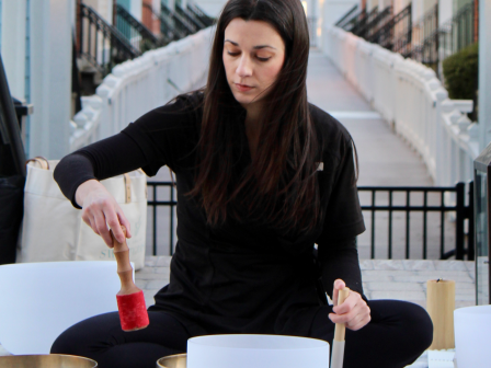 A person is sitting outside, playing singing bowls with a mallet, surrounded by various instruments and a lantern.