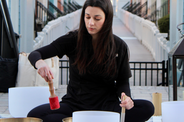 A person is sitting outside, playing singing bowls with a mallet, surrounded by various instruments and a lantern.