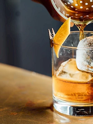A hand pours a drink from a strainer into a glass with ice and a lemon twist garnish on a bar surface.