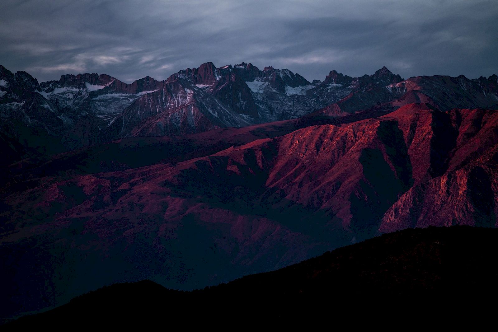 A mountain range is illuminated by dramatic reddish sunlight under a cloudy sky, casting shadows over rugged terrain.
