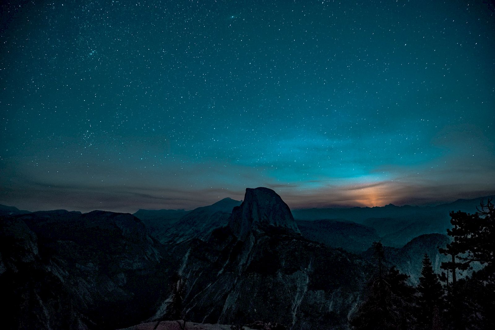 A starry night sky above a silhouetted mountain range with a subtle glow on the horizon and scattered trees in the foreground.