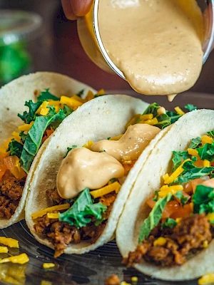 Three tacos with ground meat, cheese, lettuce, and tomato are on a plate, topped with creamy sauce being poured over them.