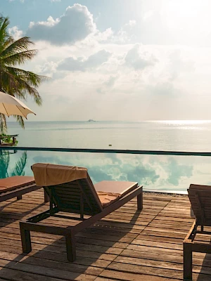 A serene beachside scene with lounge chairs, umbrellas, a pool, and palm trees overlooking the ocean under a partly cloudy sky.