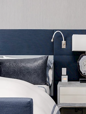 A modern bedroom with a blue headboard, white and blue bedding, a nightstand, a decorative lamp, and a mounted reading light.