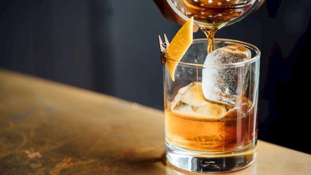 A cocktail is being poured into a glass with ice and a twist of citrus peel.