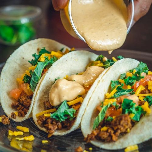 Three tacos with ground meat, cheese, lettuce, and tomatoes are being topped with a creamy sauce on a plate.