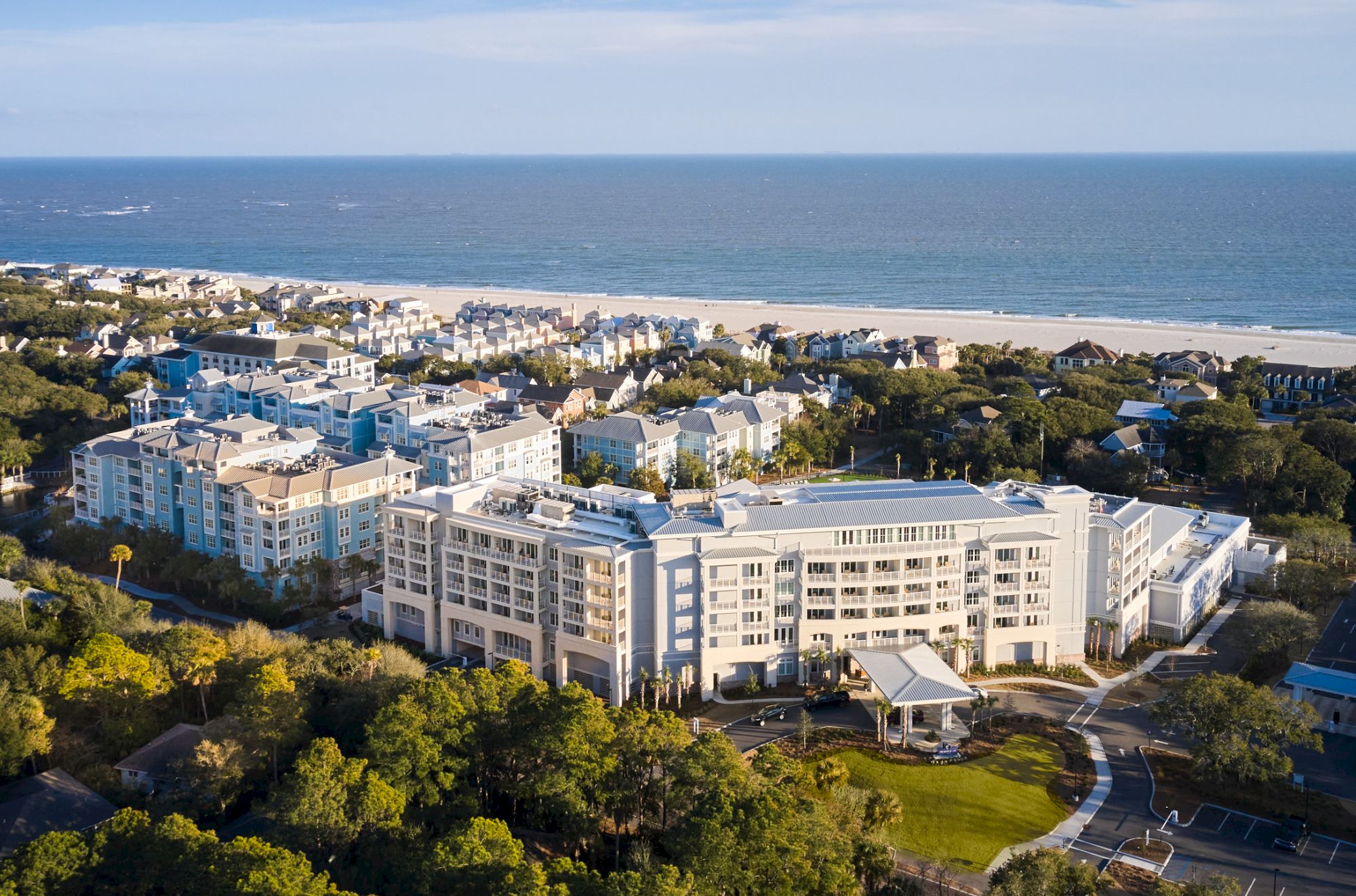 The image shows a large seaside hotel or resort surrounded by trees and residential buildings, with the ocean in the background.