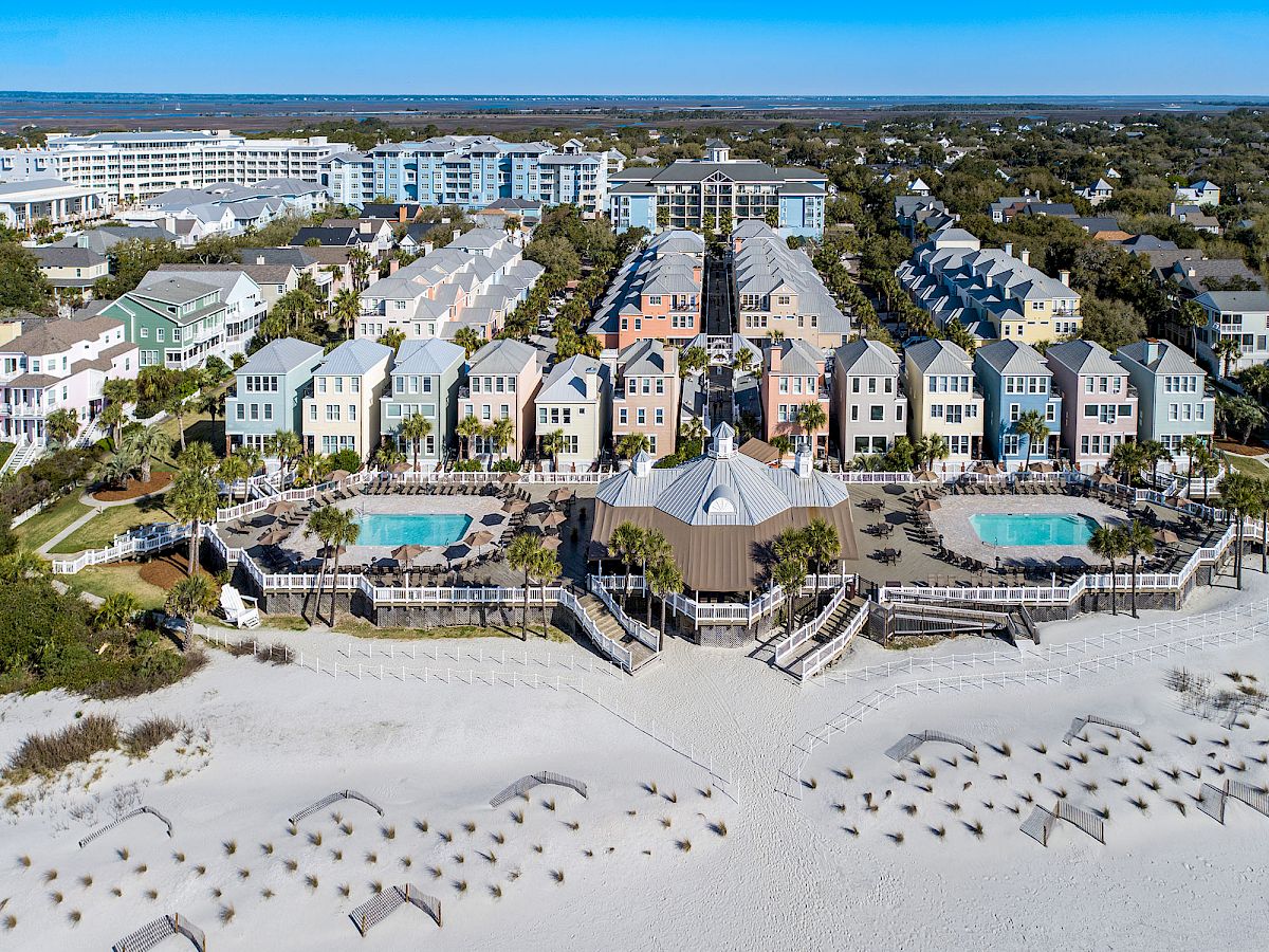 Aerial view of colorful beachfront houses, two pools, and a sandy beach with pathways and vegetation, set against a clear blue sky.