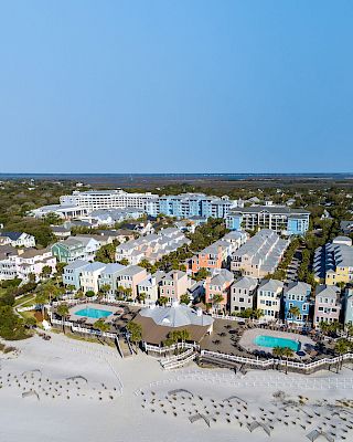 A beachfront resort with colorful houses, two pools, and sandy walkways near lush greenery and ocean views.
