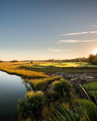 A serene landscape at sunrise with a river, marshlands, a house, and trees in the background.