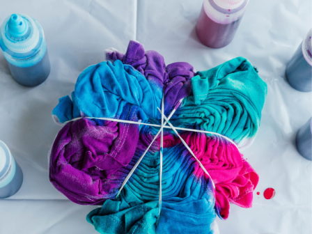 A tie-dye project is underway, with fabric bundled and dyed in blue, purple, and pink, amidst various dye bottles on a plastic sheet.