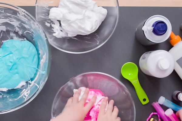 A child is mixing pink slime in a bowl, surrounded by bowls of ingredients like shaving cream, glue, and coloring tools on a table.