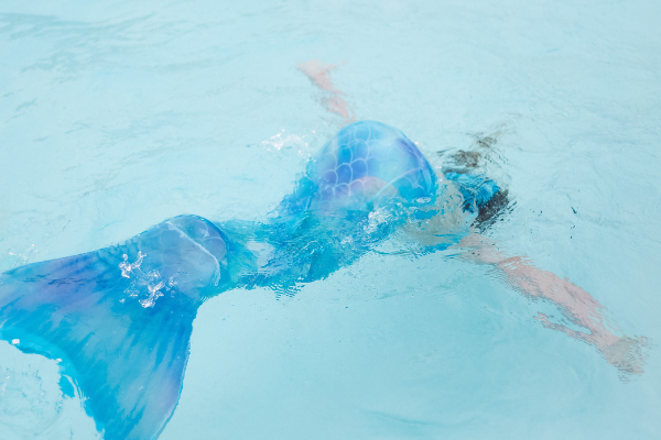 A person in a mermaid costume is swimming underwater, showing a vibrant blue and purple tail in clear water.