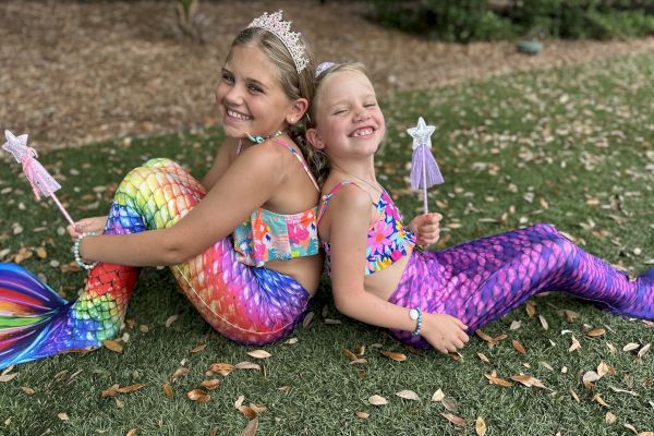 Two kids in colorful mermaid costumes and crowns, sitting back-to-back on grass, holding star wands, smiling.