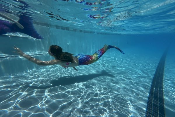 A person wearing a colorful mermaid tail swims underwater in a pool, with light reflections on the pool floor.