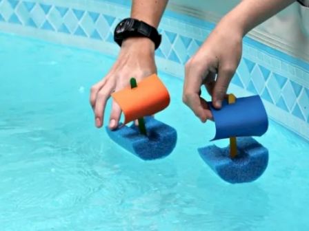 Two hands are holding small toy boats with colorful sails, made of foam, floating in a swimming pool.
