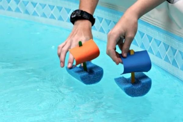 Two hands are holding small toy boats with colorful sails, made of foam, floating in a swimming pool.