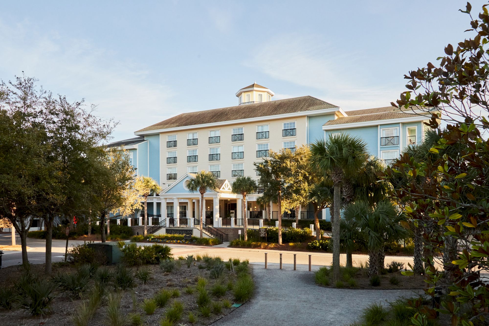 A large, light blue hotel building surrounded by trees and greenery is depicted, under a clear sky.