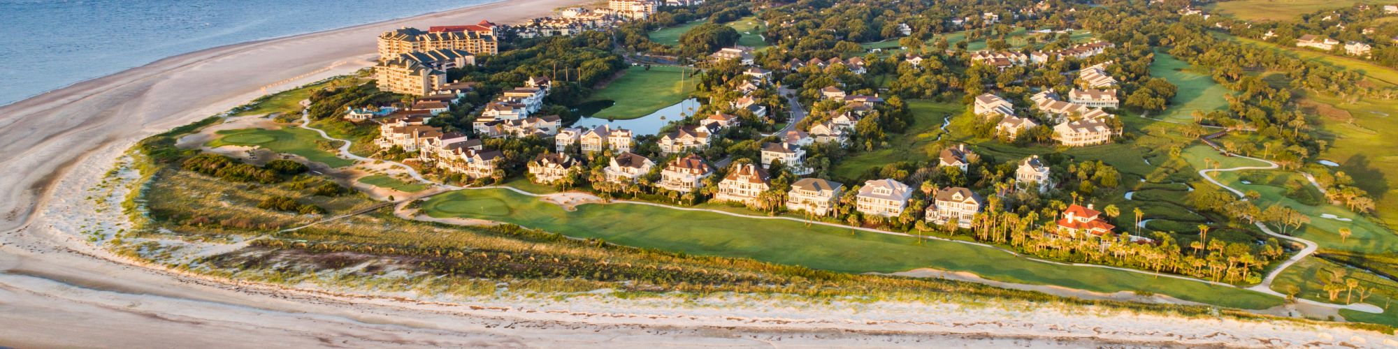 Aerial view of a coastal area with a sandy beach, ocean, and residential buildings surrounded by greenery on a peninsula.
