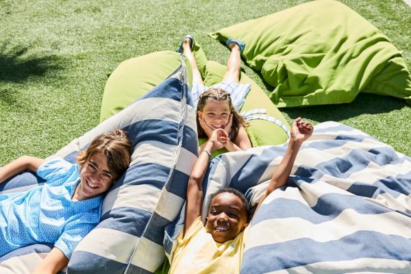 Three happy children lounging on large bean bags outdoors, smiling and enjoying the sunny day on a grassy surface.