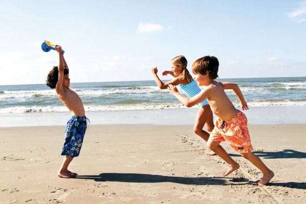 Three children playing on a sunny beach, with one holding a toy and the others running toward them, ocean waves in the background.