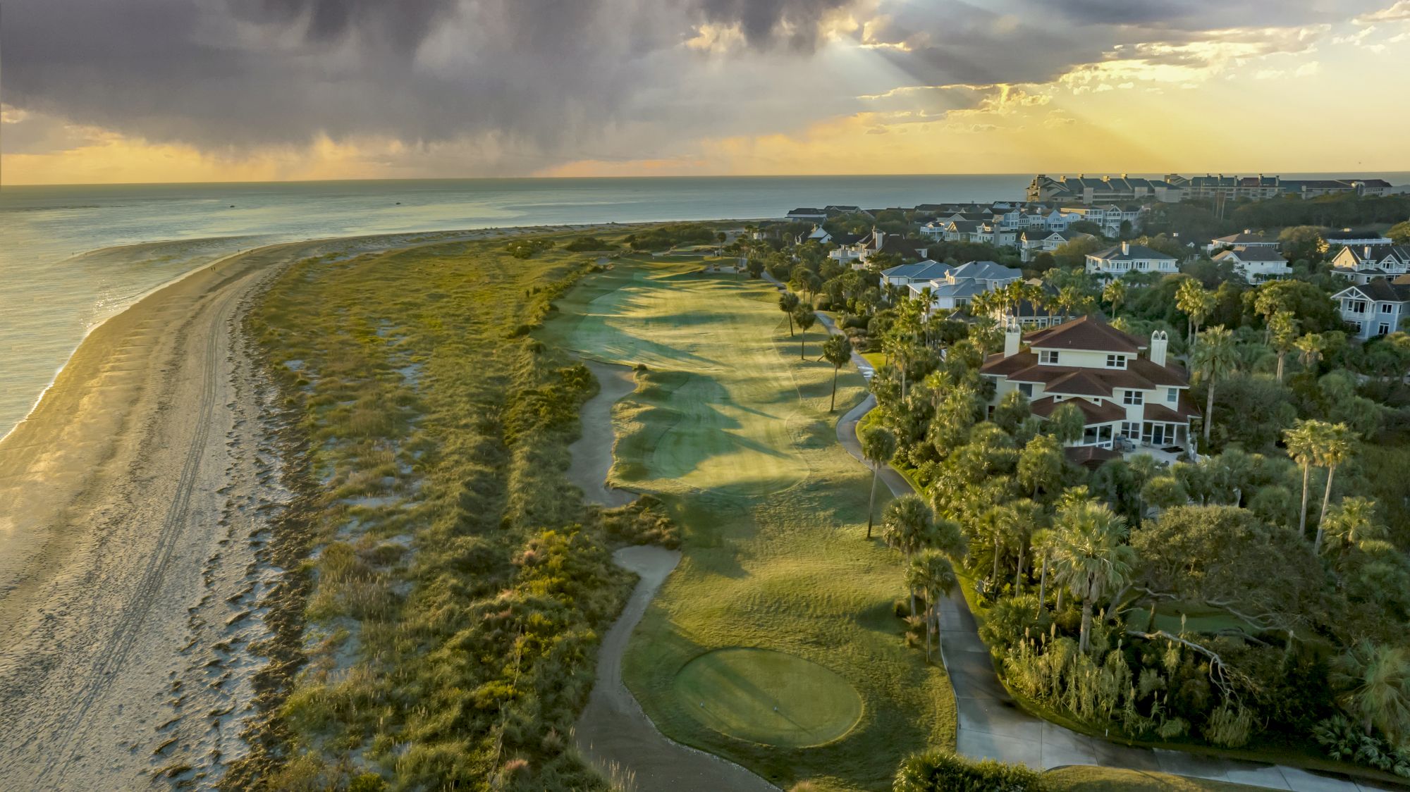 A coastal golf course with sand dunes, surrounded by lush greenery and residential houses under a dramatic sky.