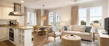 A modern, open-plan living area features a kitchen, dining table, and seating area with light furnishings and large windows.