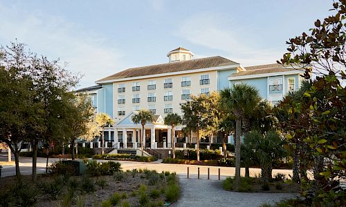 This image shows a large building with a light blue exterior, surrounded by trees and greenery, under a clear sky.