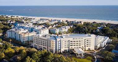 A coastal resort with several buildings, surrounded by trees, overlooking a beach and ocean under a blue sky.