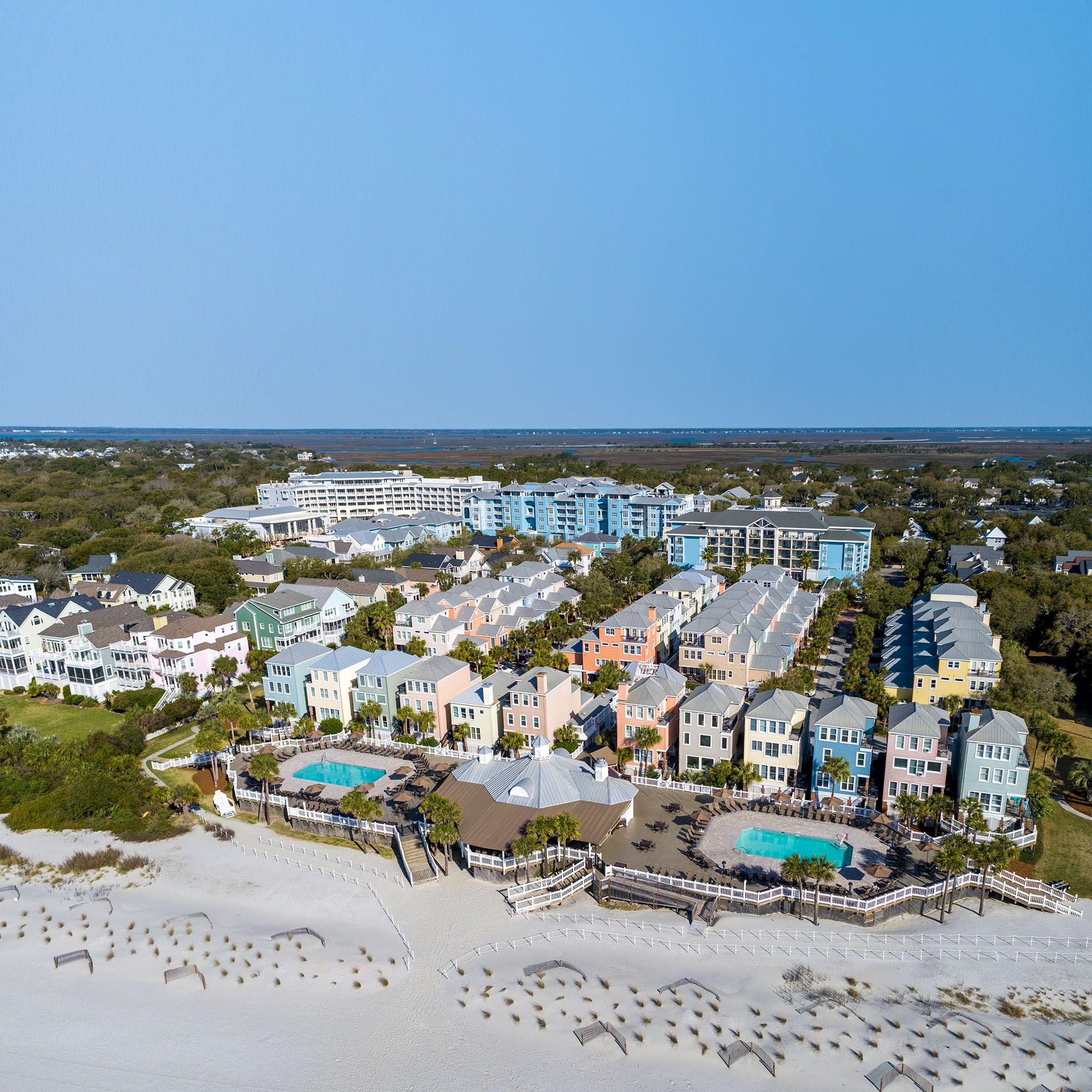 Aerial view of a colorful coastal resort with pastel houses, pools, and a sandy beach, set against a backdrop of greenery and clear skies.
