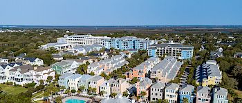 Aerial view of a coastal resort with pastel-colored buildings, two pools, and beach umbrellas on the sandy shore, under a clear blue sky.