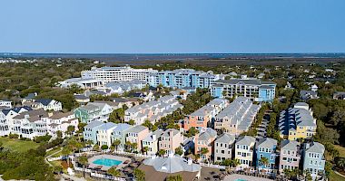 Aerial view of a coastal resort with pastel-colored buildings, two pools, and beach umbrellas on the sandy shore, under a clear blue sky.